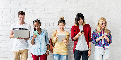 5 students lined up to a brick wall looking at their phone, laptop or tablet