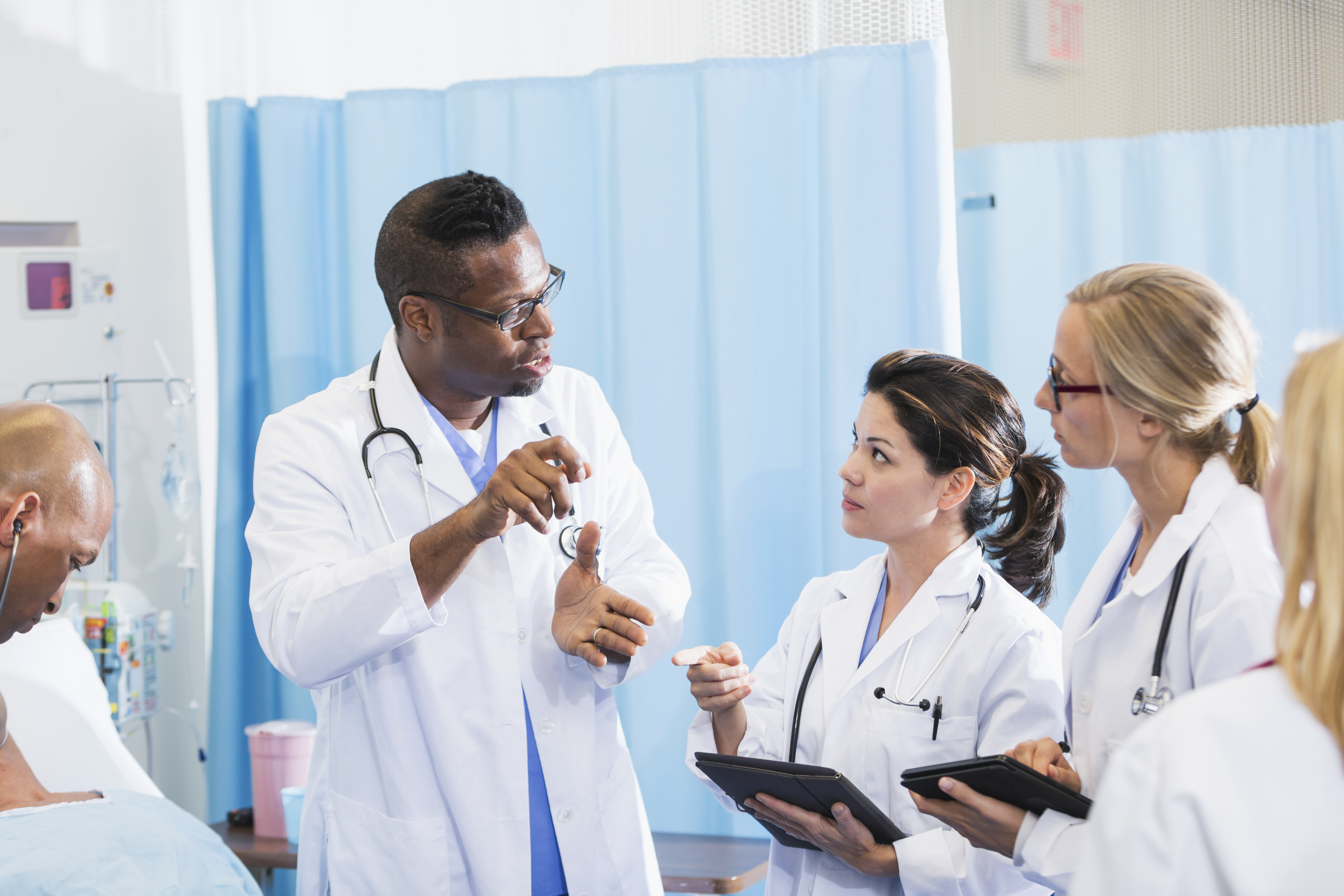 A group of multi-ethnic doctors in a hospital room, wearing white lab coats, having a discussion.  Two of the female doctors are holding digital tablets.