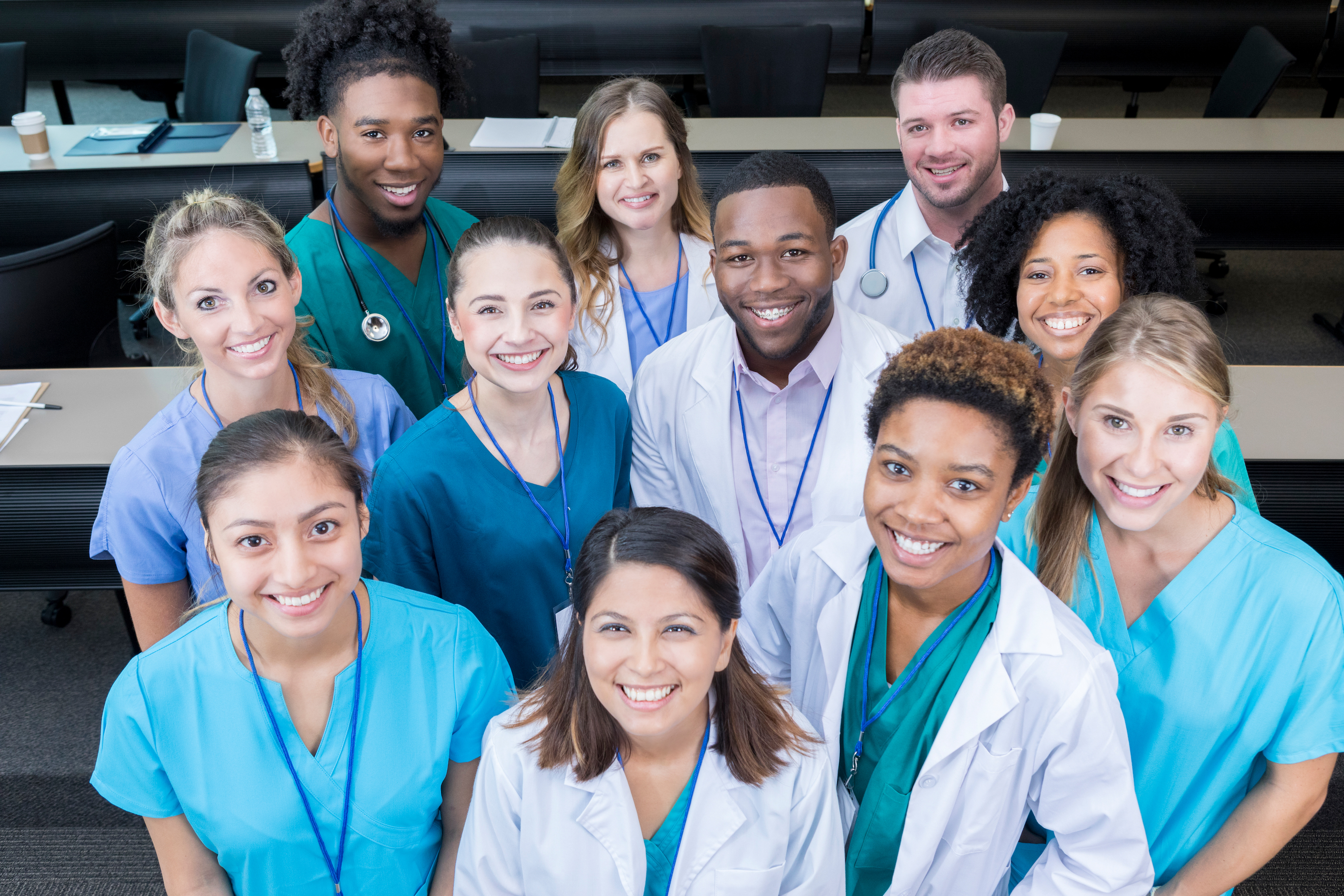 A group of medical students stand in their classroom.  They look up and smile for the camera.
