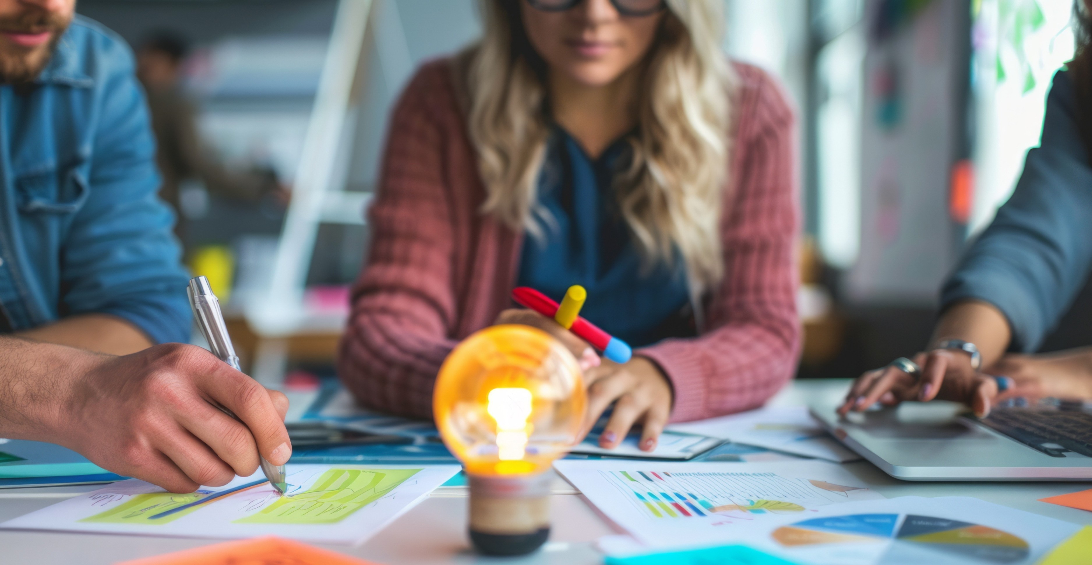 A team collaborates around a table filled with charts, sticky notes, and diagrams. A glowing light bulb sits at the center, symbolizing ideas. One person writes with a pen, another types on a laptop, and a third holds colorful markers.