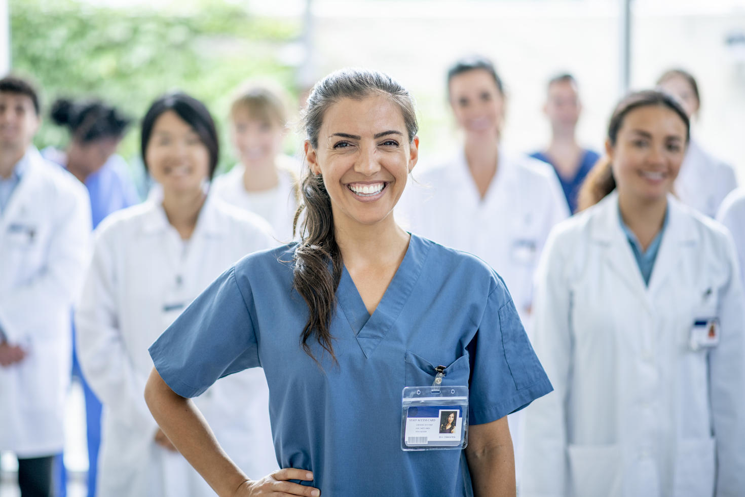 A female medical student of Hispanic ethnicity smiles for the camera as she stands in front of her group of medical professionals in the background.