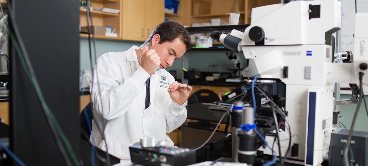A person in a lab coat examines a sample through a microscope in a laboratory.