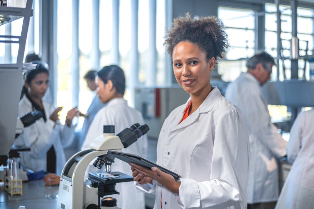 Researcher in a laboratory holding a tablet beside a microscope, with colleagues working in the background.