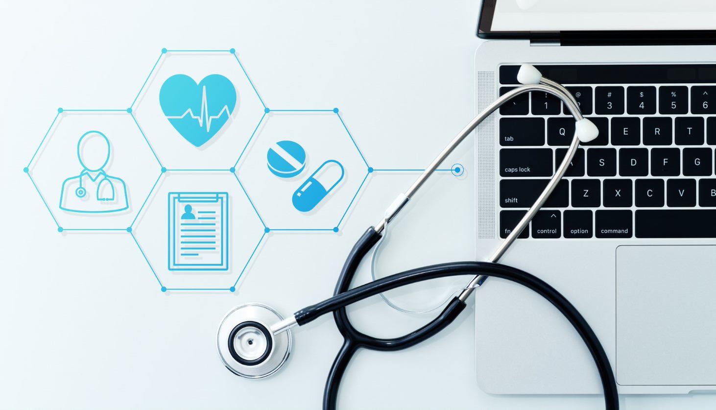 Top-down view of a black stethoscope placed in front of a laptop. Blue medical icons, including a heart, a medical cross, and other healthcare symbols, appear on the screen.