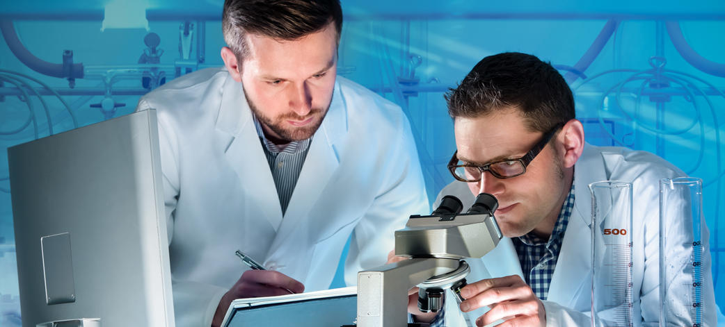 A male researcher is looking in a microscope while another male researcher writes notes in a pad