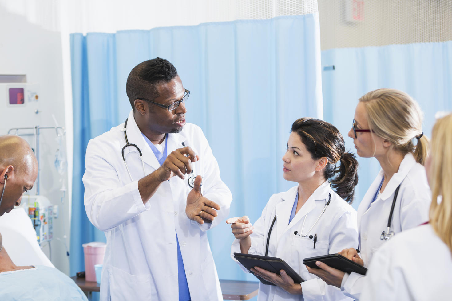 A group of multi-ethnic doctors in a hospital room, wearing white lab coats, having a discussion. Two of the female doctors are holding digital tablets.