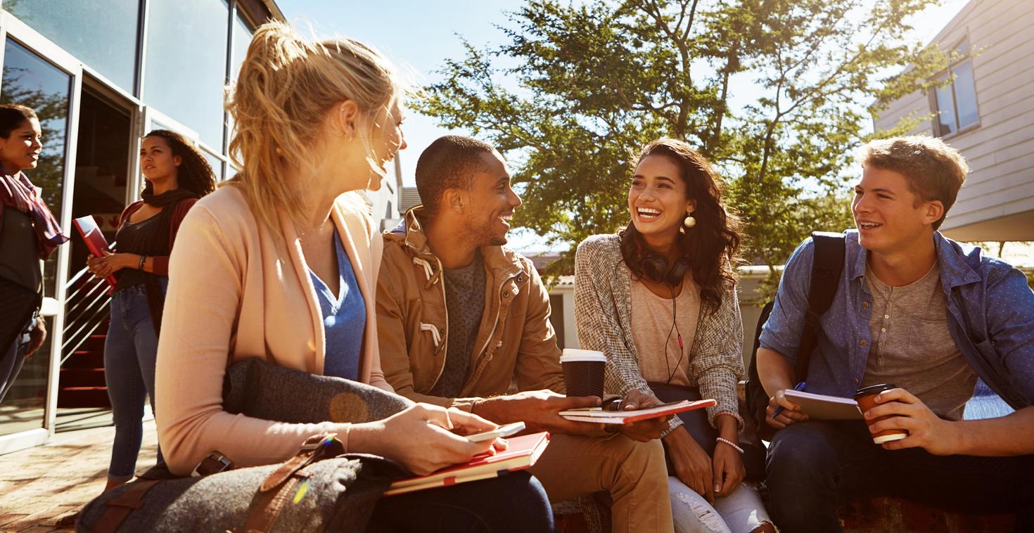 A group of four people sit outdoors, chatting and smiling under a sunlit tree