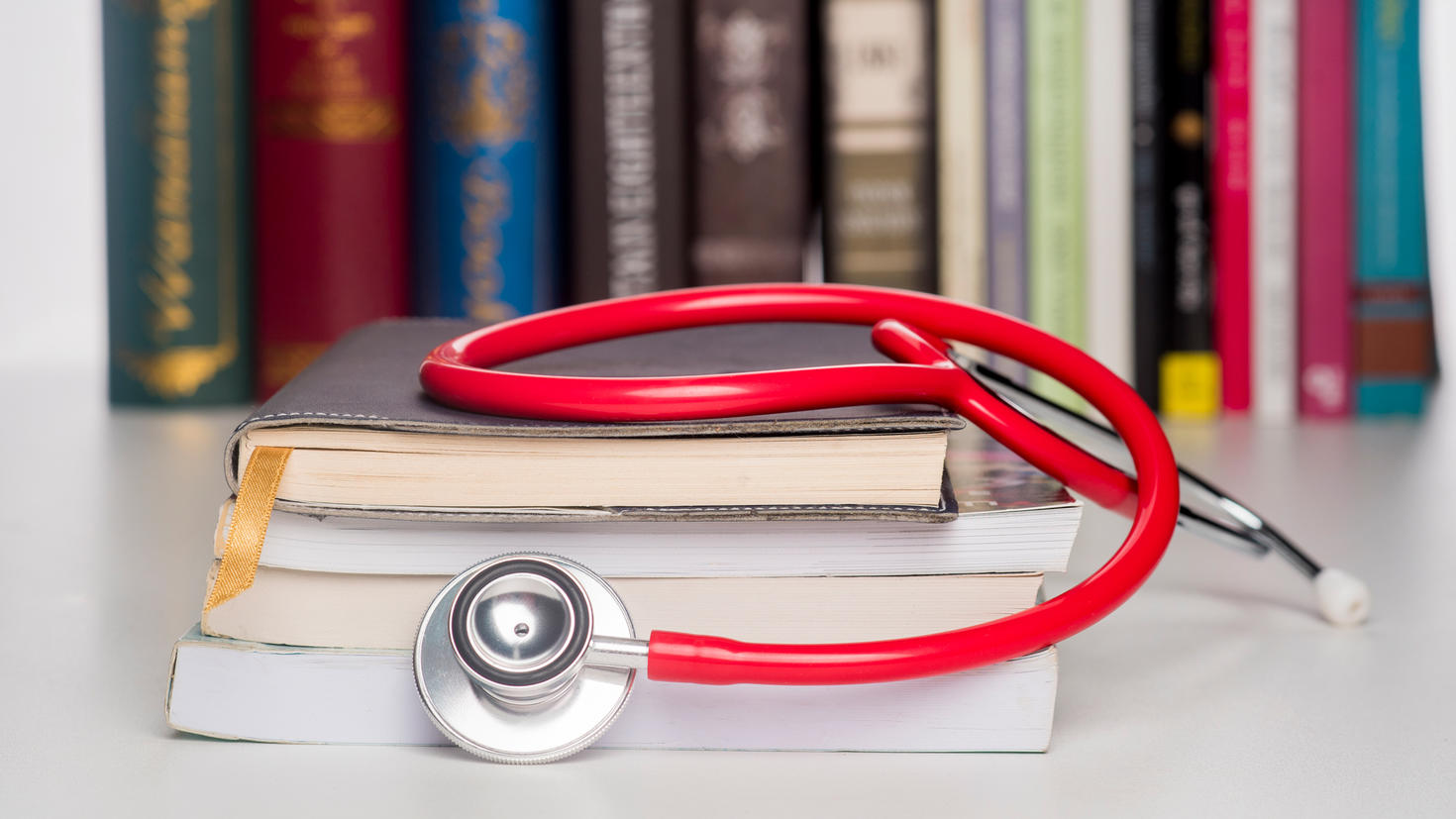 Close-up of a red stethoscope resting on a stack of books in front of a row of books in a library.