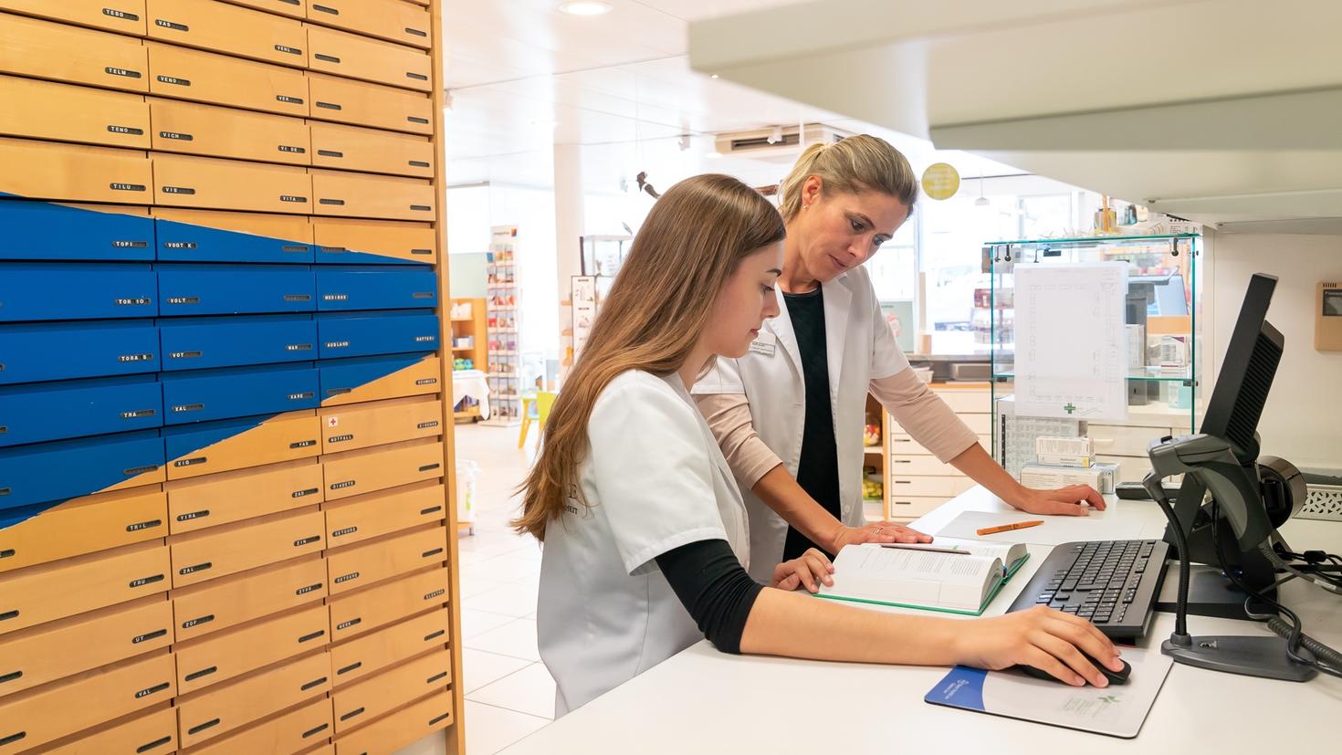 A female pharmacist helps and assists her young aprrentice trainee during studies of medical books