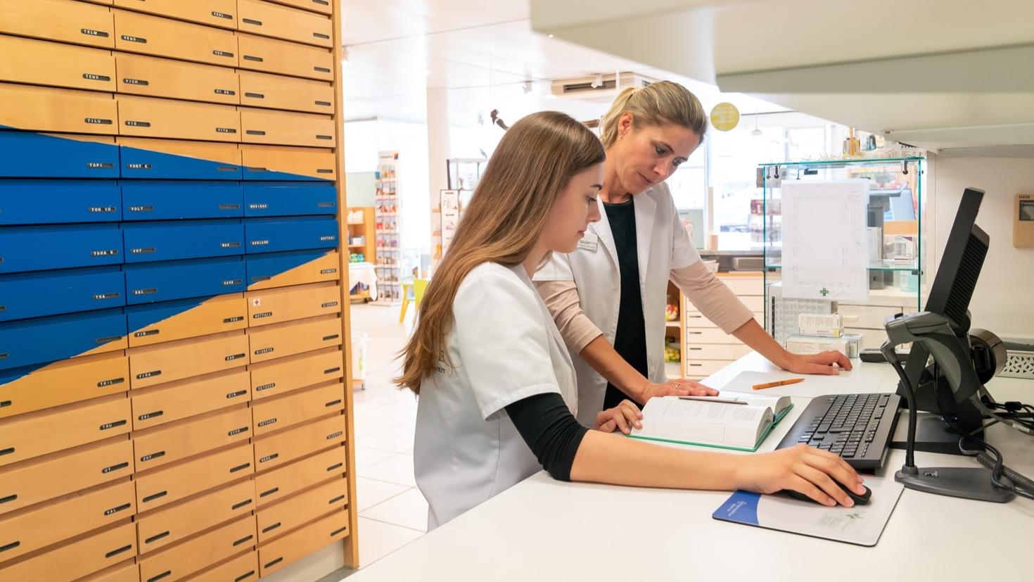 Female blonde pharmacist helps and assists her young aprrentice trainee during studies of medical books
