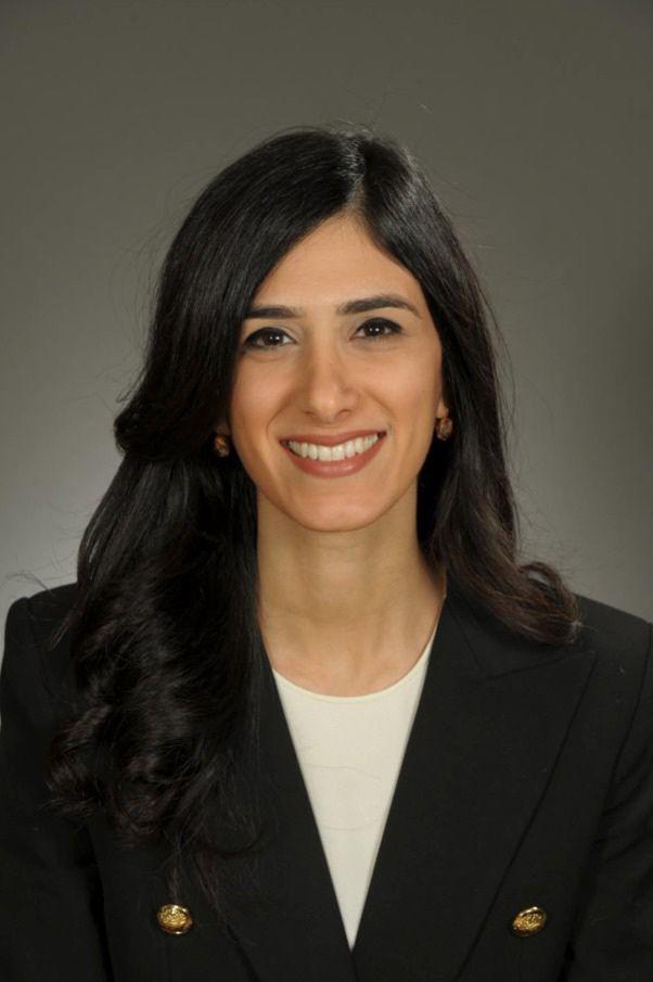 Dr. Elise Azzi smiling while wearing a black blazer with brass buttons and a white blouse over a cool taupe grey background