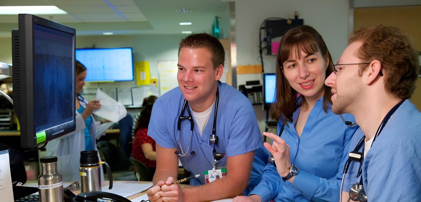 Three medical professionals in blue scrubs, with stethoscopes, are engaged in a discussion around a computer screen in a busy hospital environment.