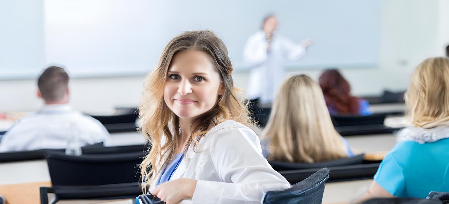 A woman in a white coat smiles and looks at the camera in a classroom setting. In the background, a person gestures while giving a lecture.
