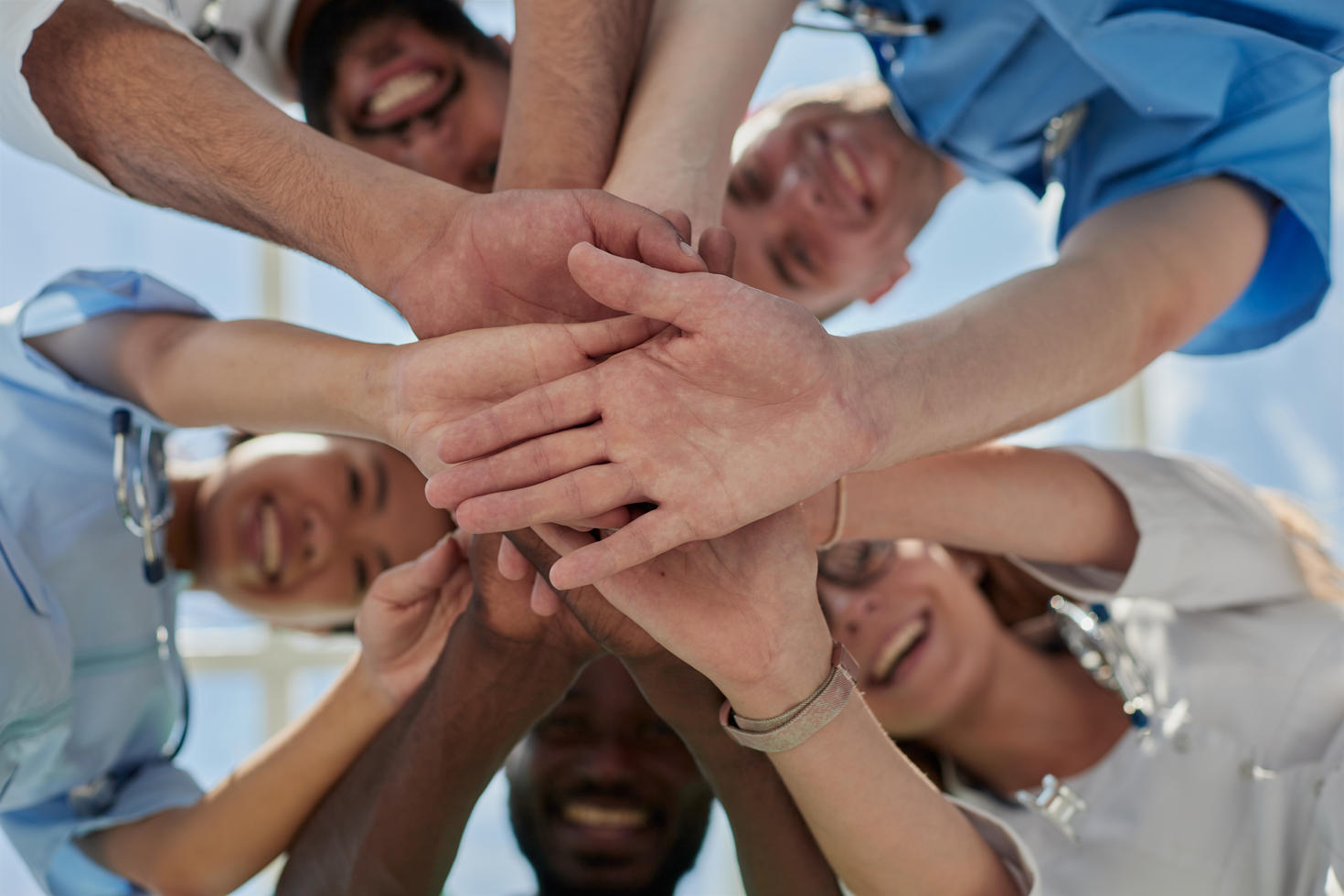 A group of individuals standing in a circle, smiling, and stacking hands in unity against a blue sky backdrop