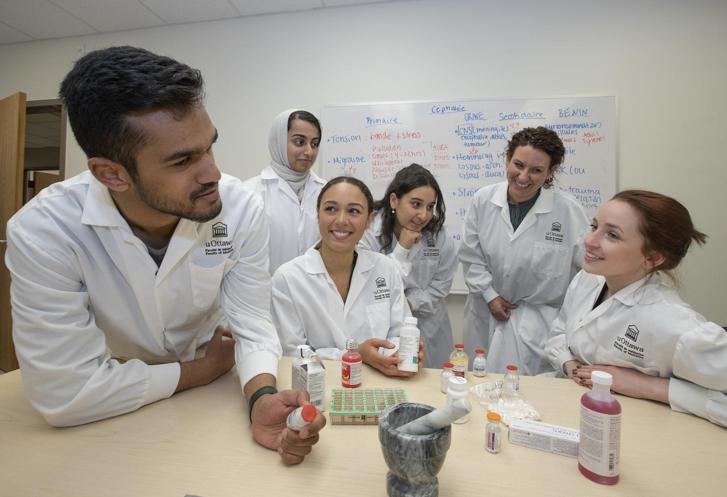 Six students in white lab coats are working together around a table, examining vials and pharmaceutical supplies in a hands-on learning environment.