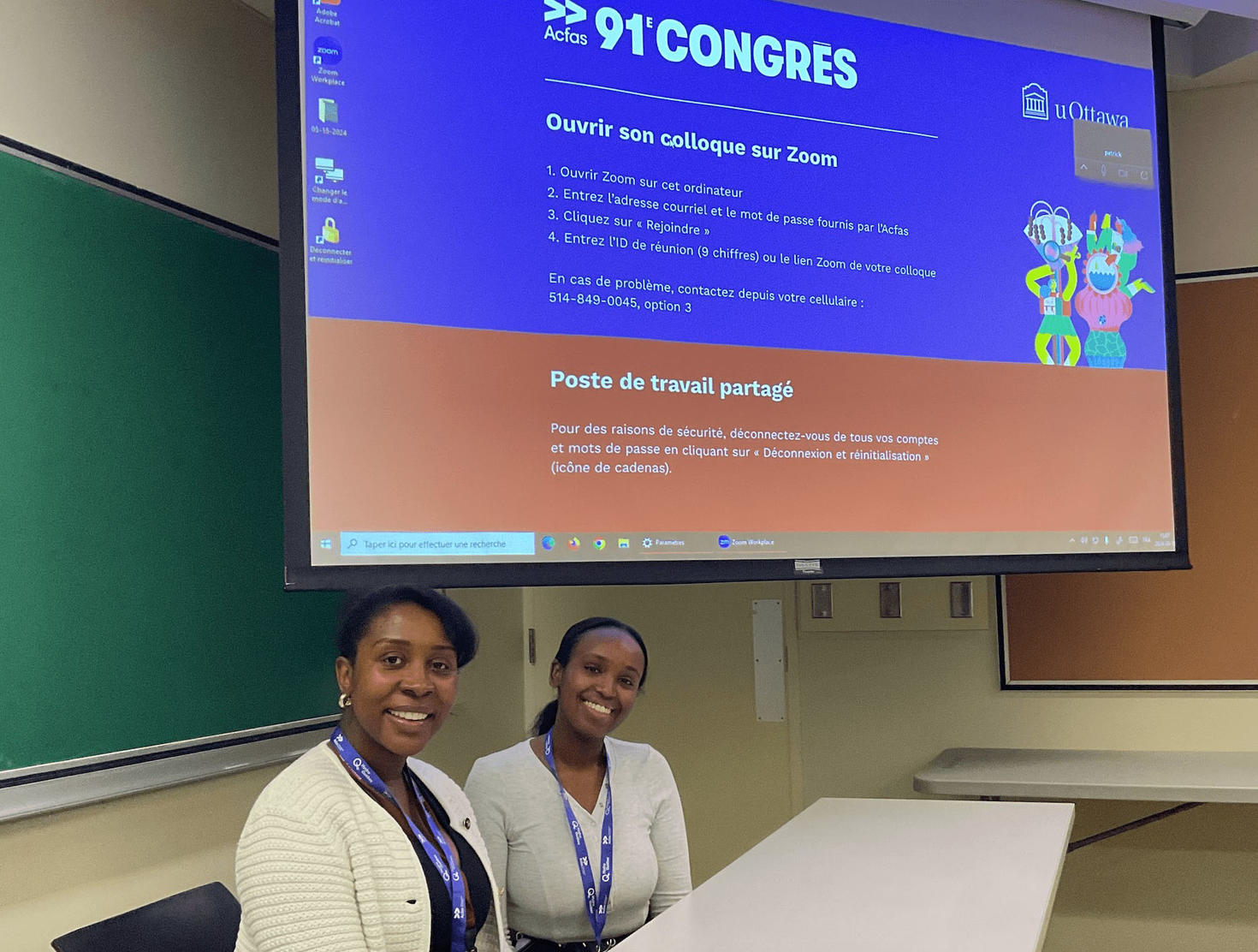 a professor and a student teacher sitting together at a table in front of a presentation screen, smiling at the camera.