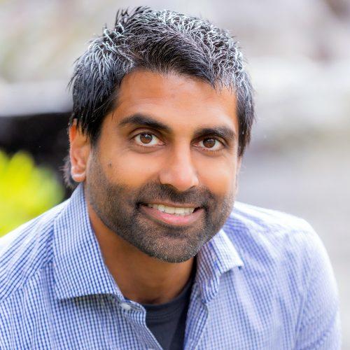 Dr. Osmaan Sheikh smiling while wearing a stripe pattern shirt and dark undershirt over a blurred background