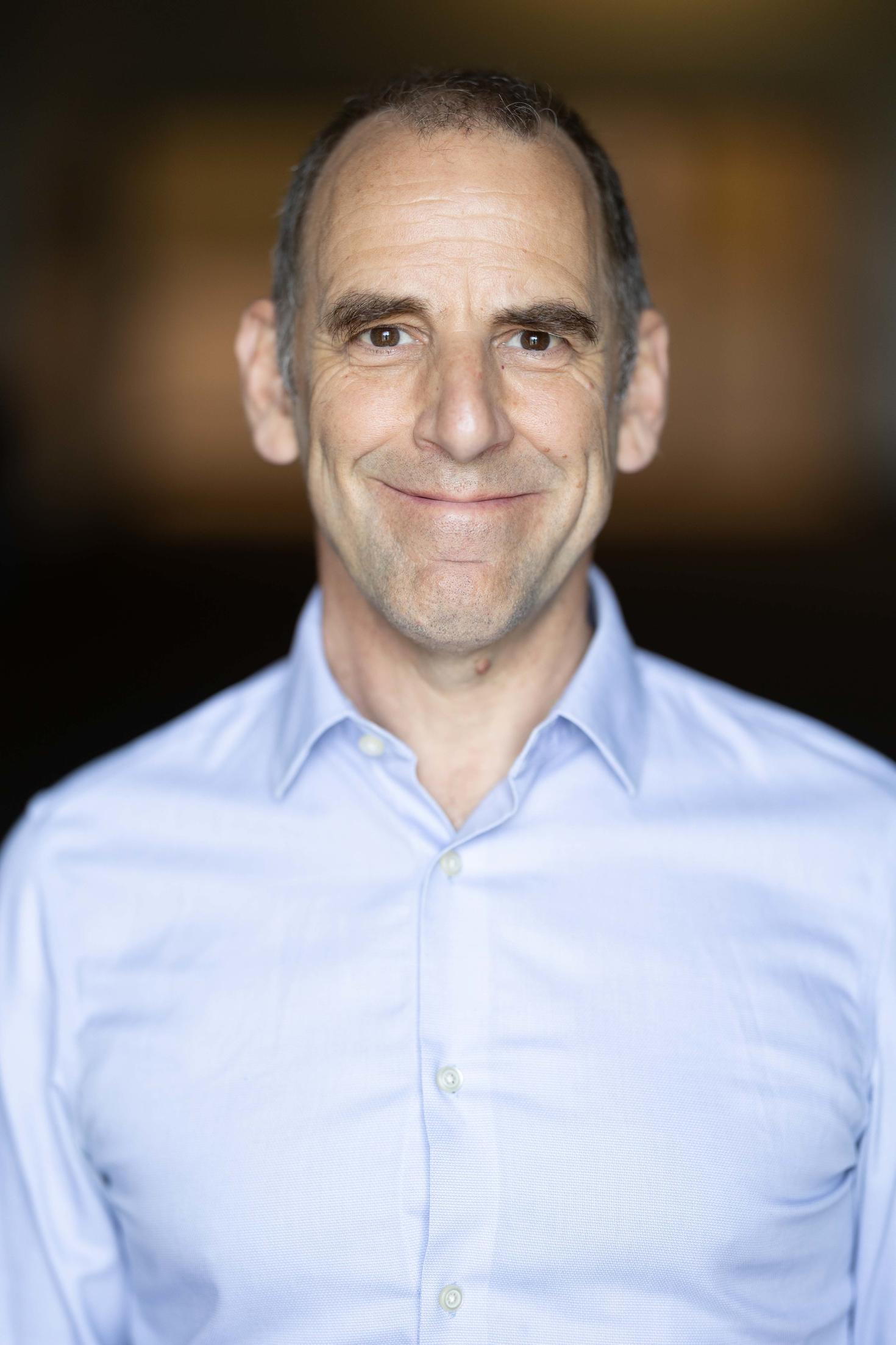 Dr. Douglas Manuel smiles at the camera, wearing a light blue button-up shirt against a softly blurred dark background.