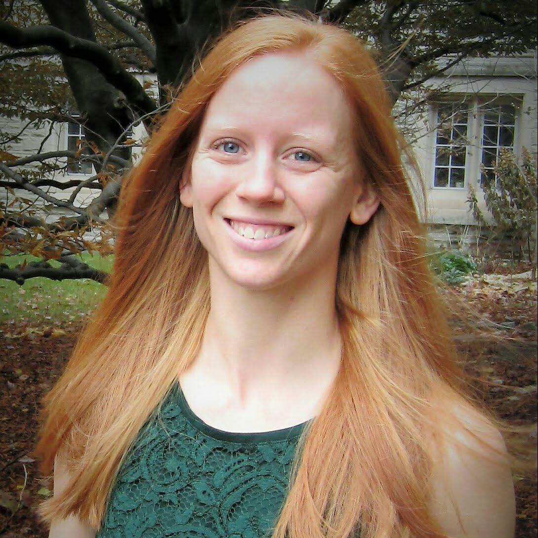 Dr. Janet Barber smiling into the camera and wearing a dark green floral pattern shirt. The background is a scenic forest.