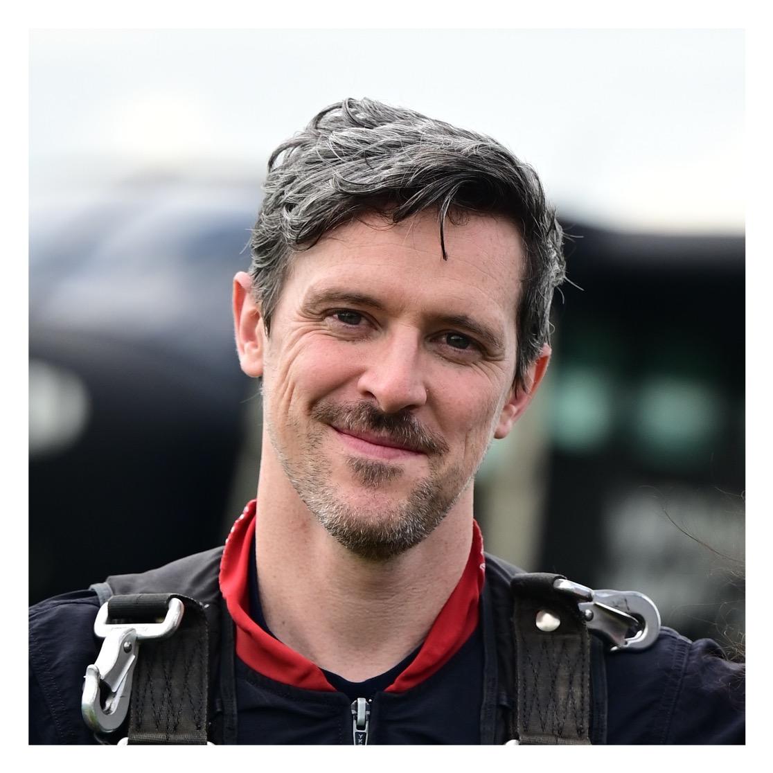 Dr. Elliott Lee smiles at the camera while wearing a dark jacket with a red collar and black shoulder straps with metal clips.