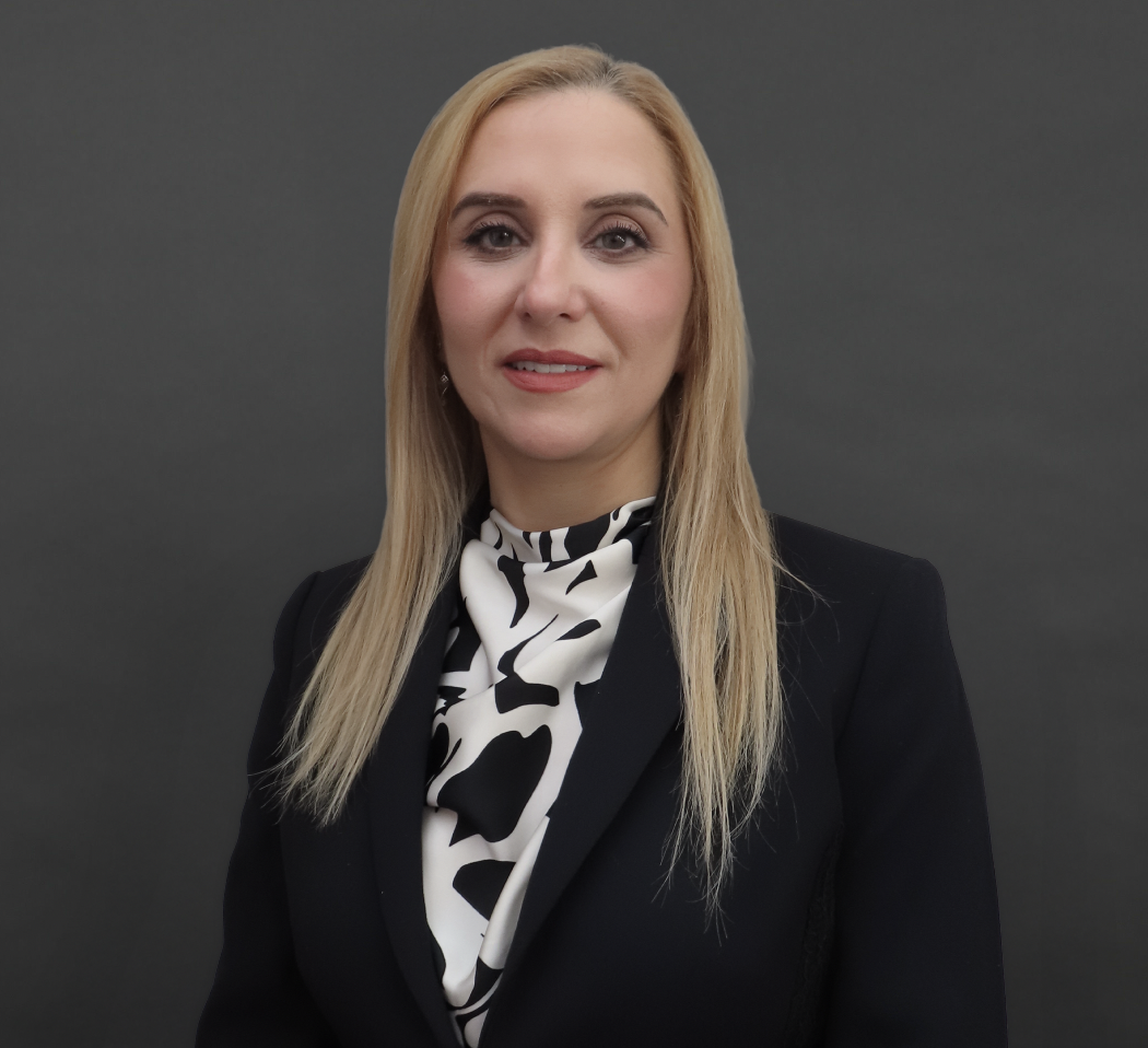 Dr. Kay-Anne Haykal looks at the camera, wearing a black blazer over a black-and-white patterned blouse against a dark grey background.