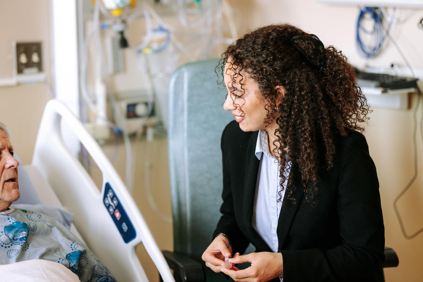 A pharmacist in a dark blazer smiles as she speaks with an elderly patient resting in a hospital bed, holding a medication cup in her hands.