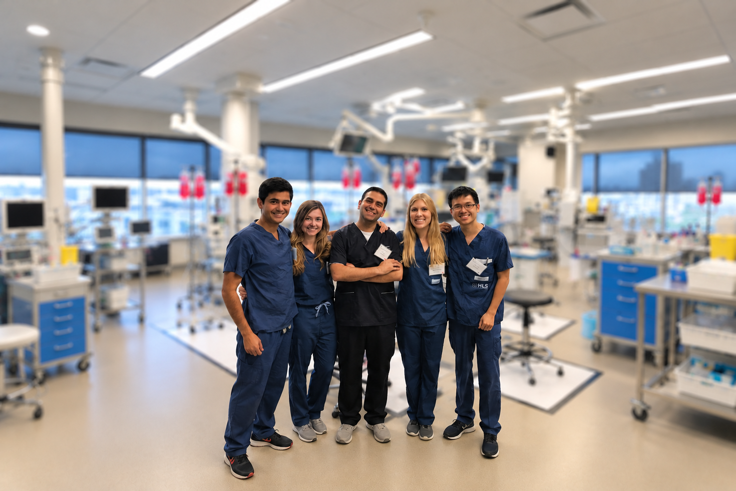 Five smiling healthcare professionals wearing blue medical scrubs pose together in a hospital room equipped with medical equipment.