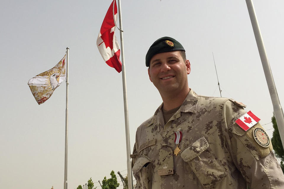 Dr. Maxime Britt-Côté standing in front of flags wearing a beige camouflage uniform.