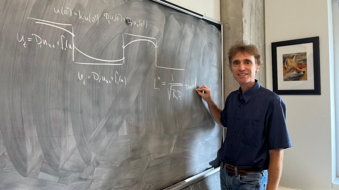 Professor Frithjof Lutscher is standing to the right of a chalkboard on which he is writing a mathematical formula.