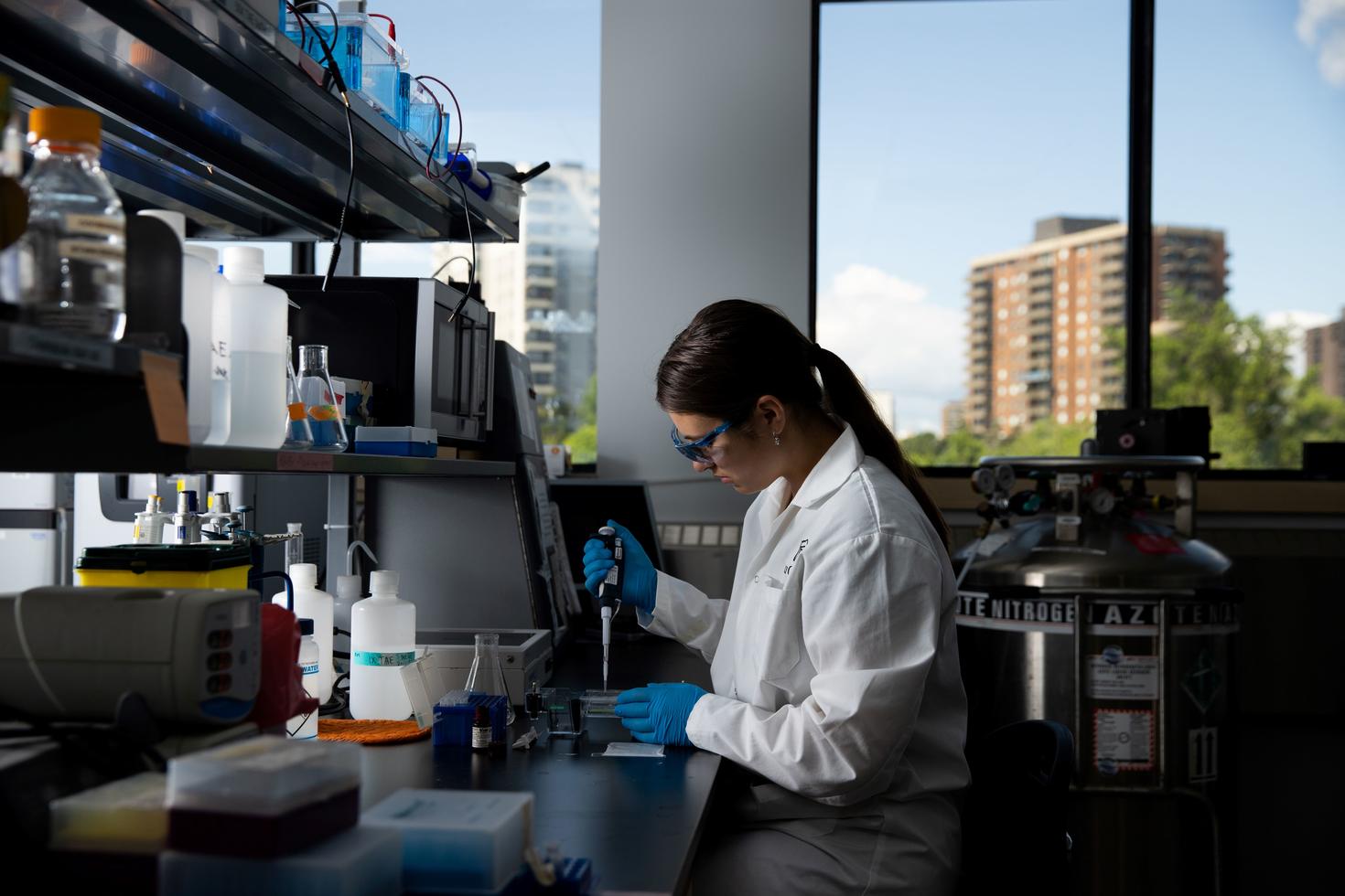 A woman wearing safety glasses and a white lab coat, sitting at a laboratory bench and holding a micropipette.