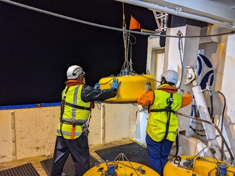 Two workers in safety vests handling and preparing to deploy a yellow ocean-bottom seismometer
