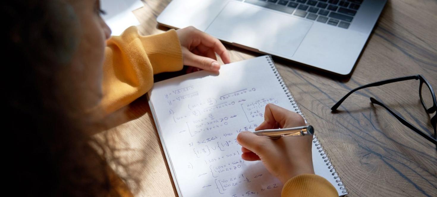 Female student wear headphones studying from home writing in workbook solving equations learning math sits at desk