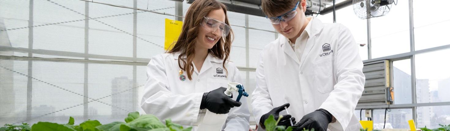 Two students in lab coats working with plants in a greenhouse