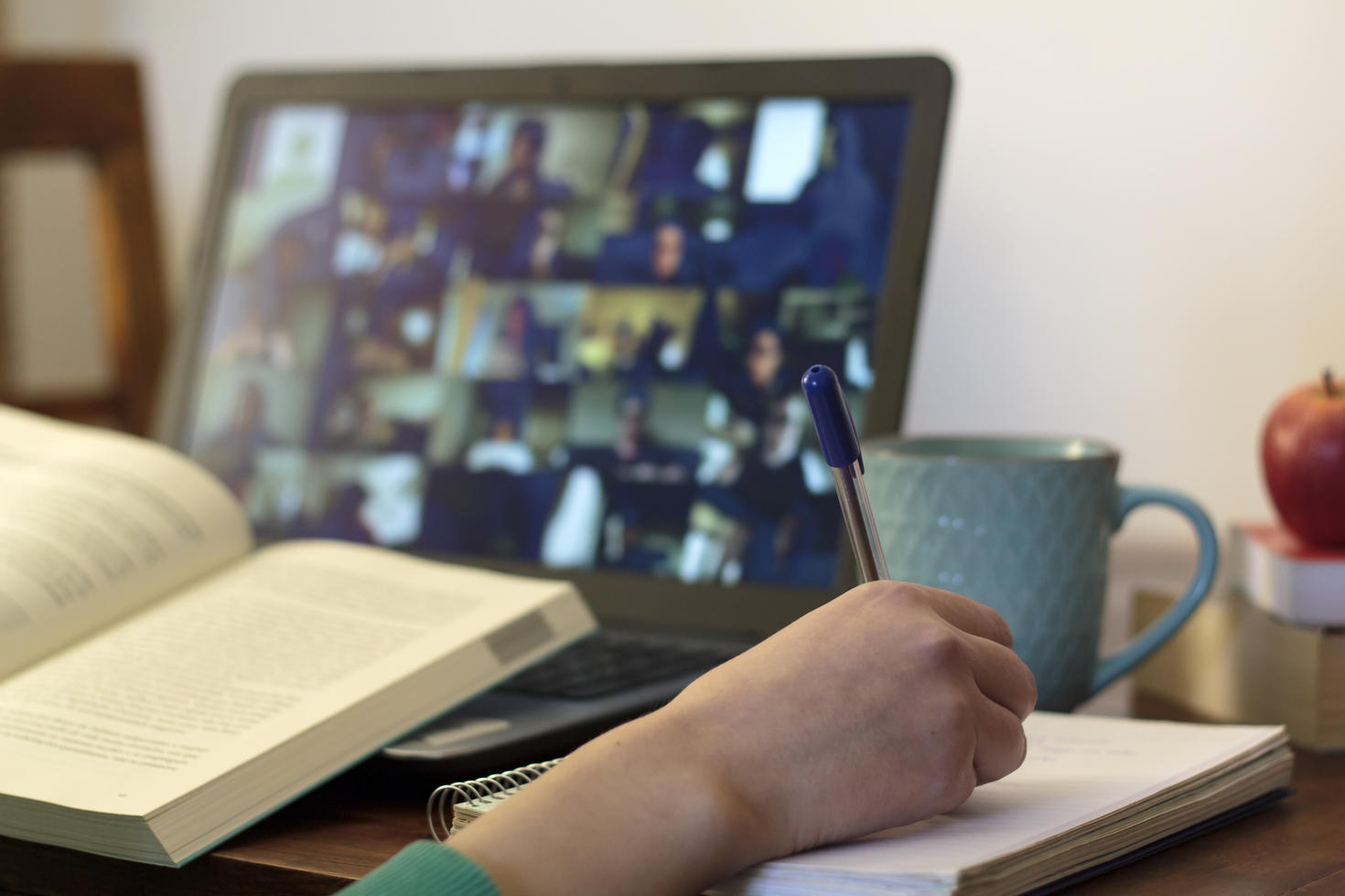 open book in front of laptop with a hand holding a pen and mug