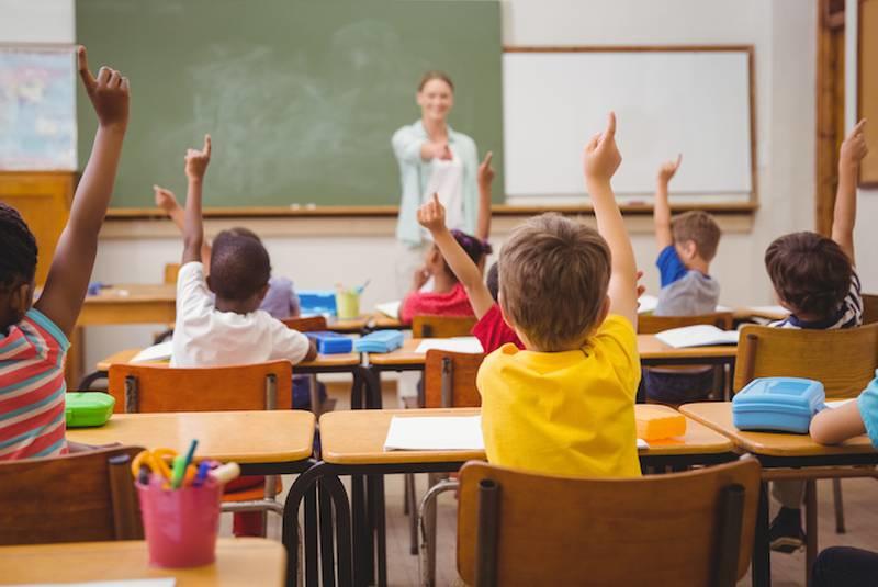 children holding up arm in classroom with teacher pointing at them