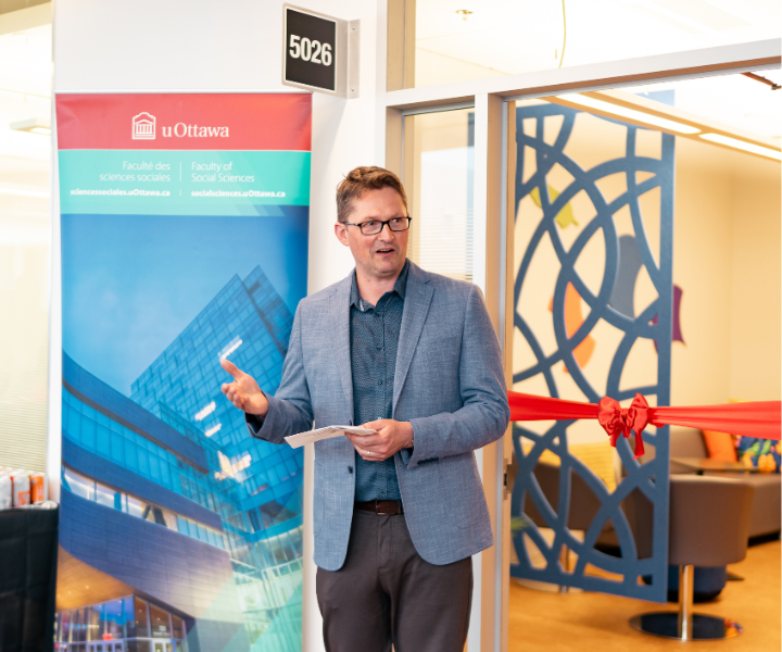 A faculty leader delivers remarks during the opening ceremony while holding prepared notes. The entrance to the affinity space, featuring decorative blue artwork and the red ribbon from the ribbon-cutting ceremony, is visible behind them.