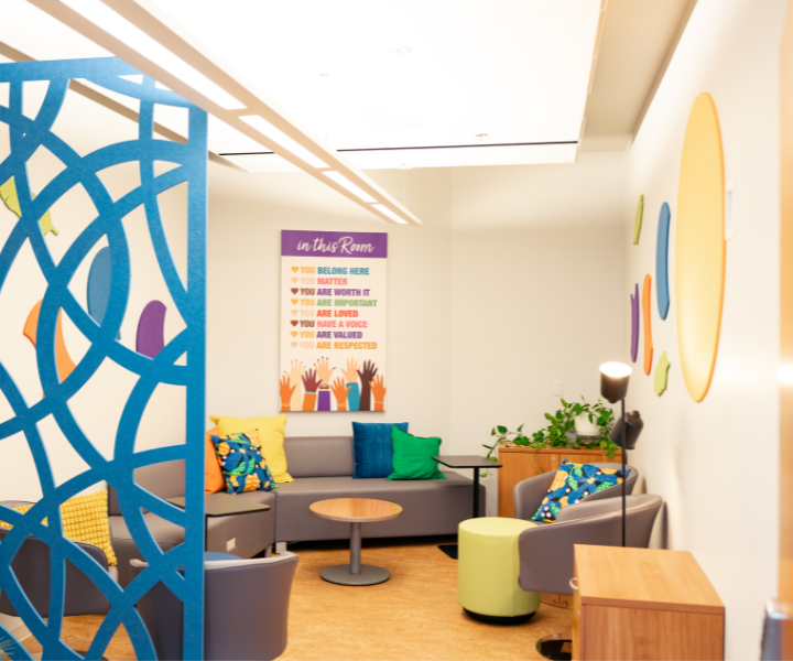A welcoming lounge area with modern seating, colourful cushions, plants, and decorative wall art. A large poster on the back wall displays affirming messages including "You belong here," "You matter," and "You have a voice," creating an inviting and inclusive atmosphere.