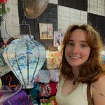 White girl with brown hair in a shop with her head close to an exotic lamp. Black board in the background with documents pinned to it