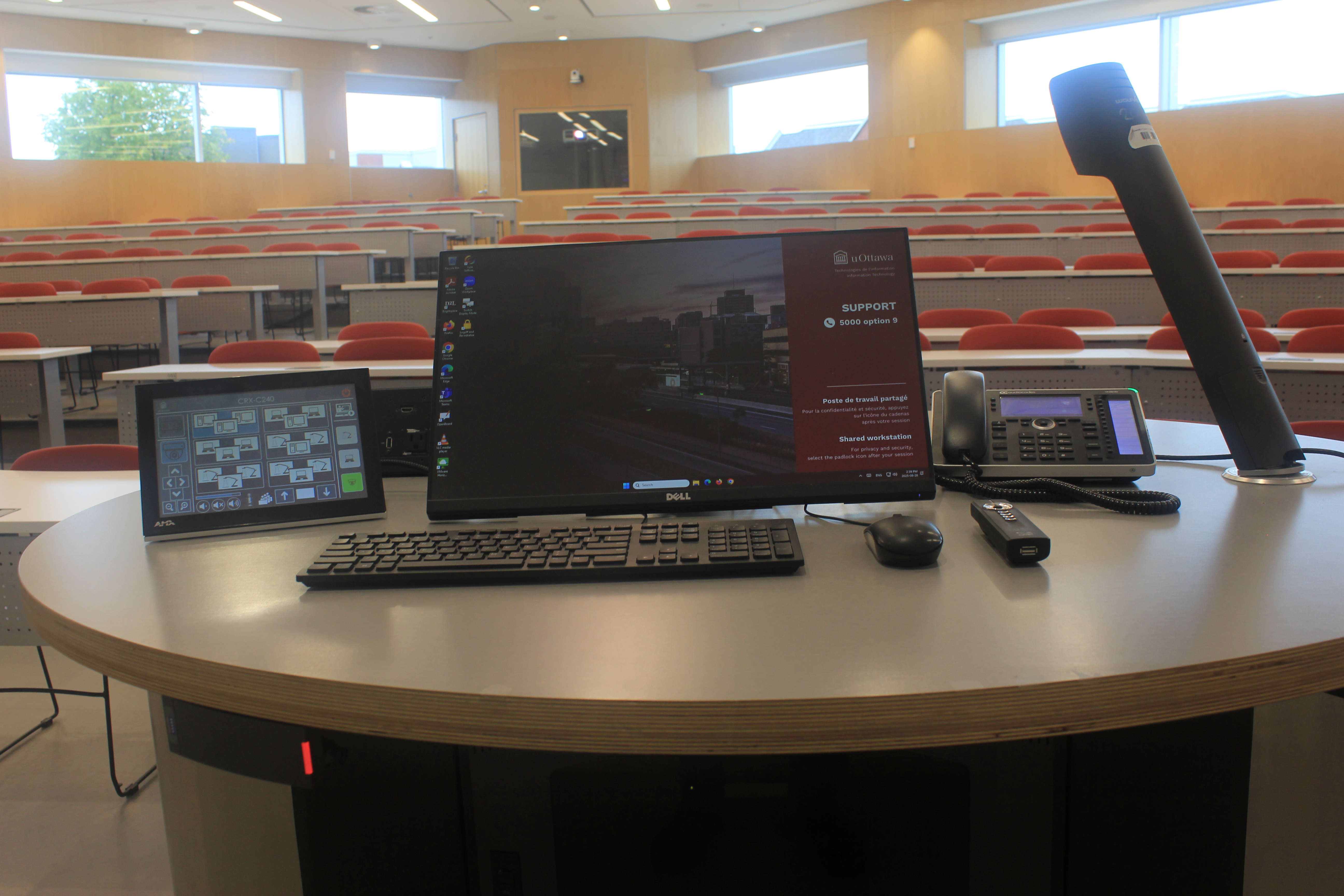 Une salle de classe avec des sièges rouges et un podium équipé d’un ordinateur, d’un microphone et d’un écran de projection.