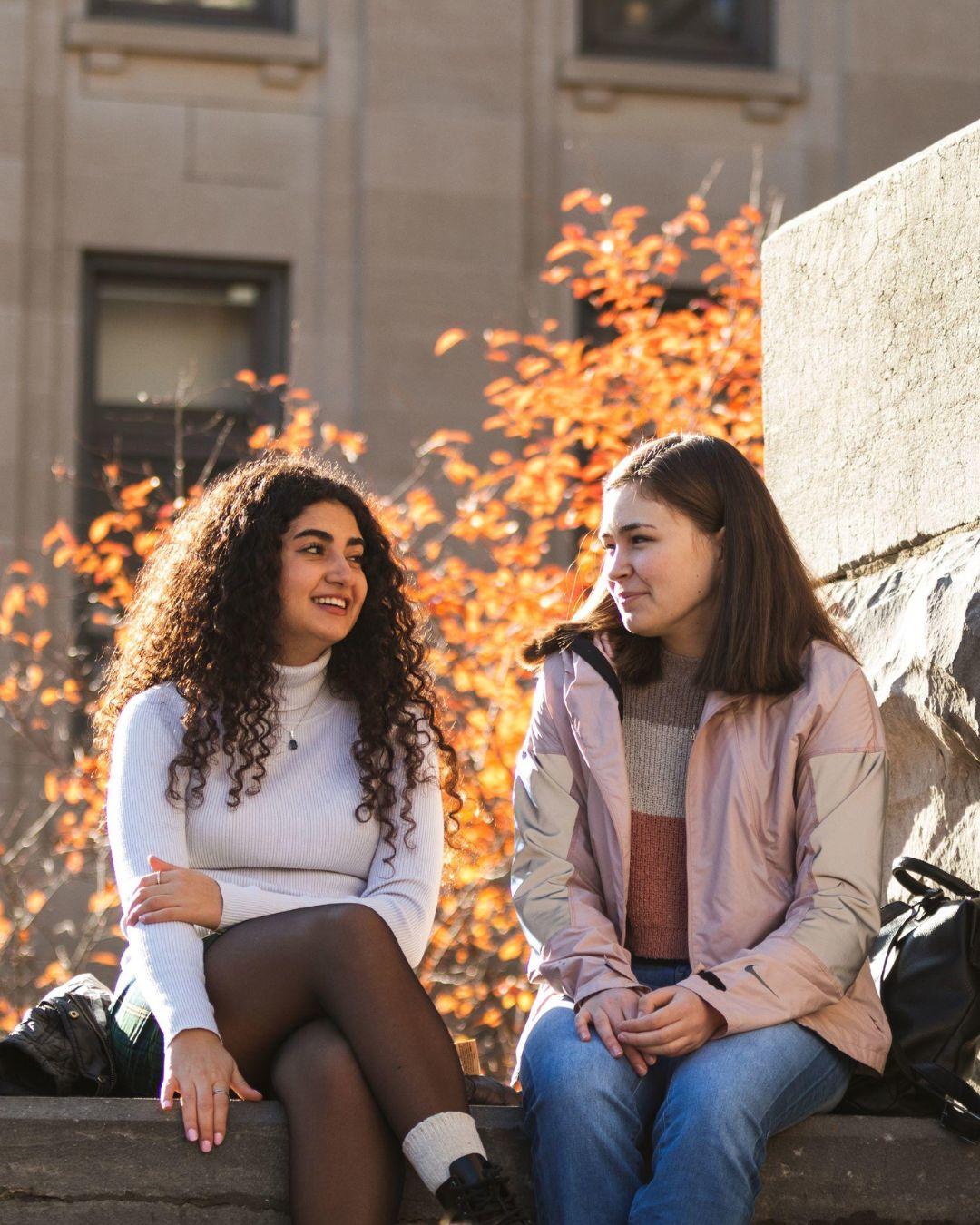 Deux étudiants assis côte à côte sur une surface de pierre. Arbres d’automne en arrière-plan.