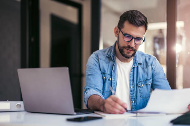 Personne assise à un bureau, travaillant avec des papiers, un ordinateur portable ouvert et un téléphone dans un bureau vitré.