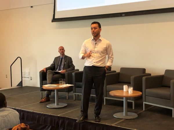 Olivier Choinière giving a presentation, Marc Morin sits in a chair in the background
