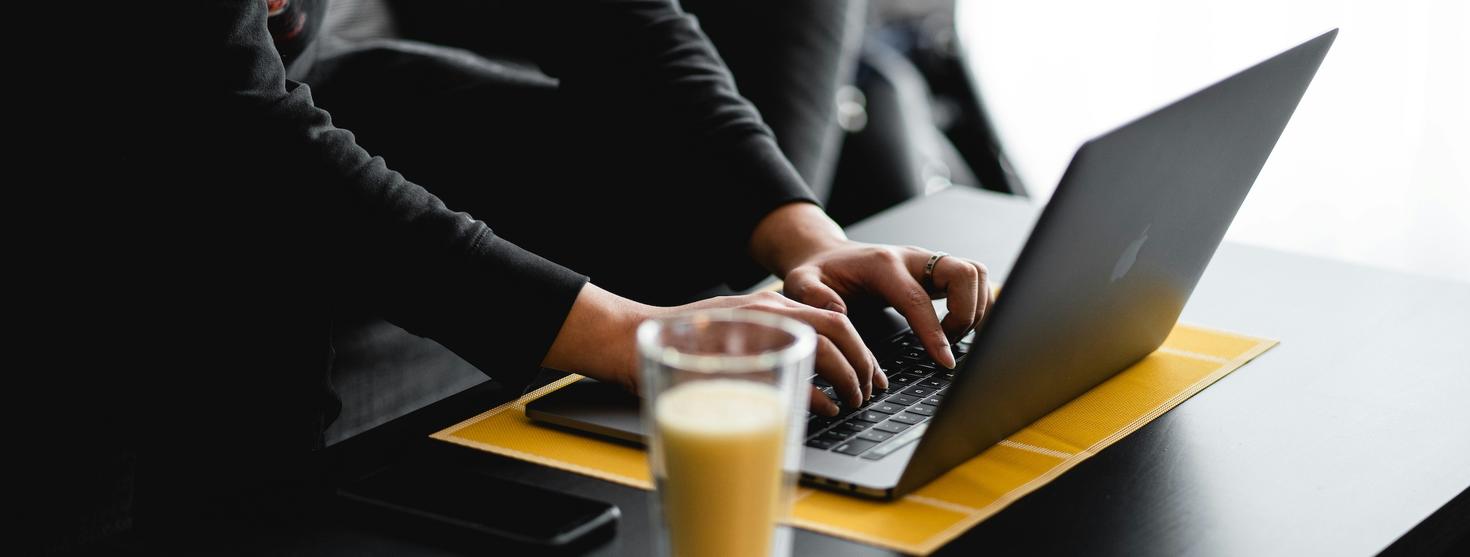 Personne écrivant sur un ordinateur portable à une table avec une boisson au premier plan