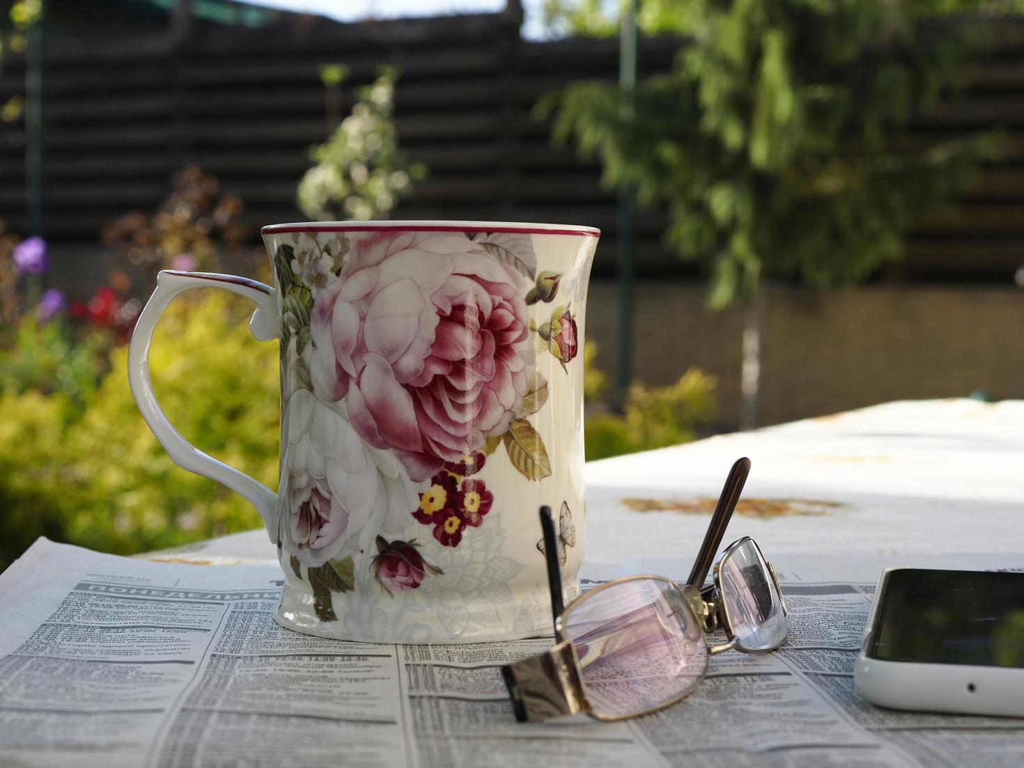 Une tasse à motif floral est posée sur une table recouverte de journaux, à l’extérieur, à côté d’une paire de lunettes et d’un smartphone, avec un jardin flou en arrière-plan.