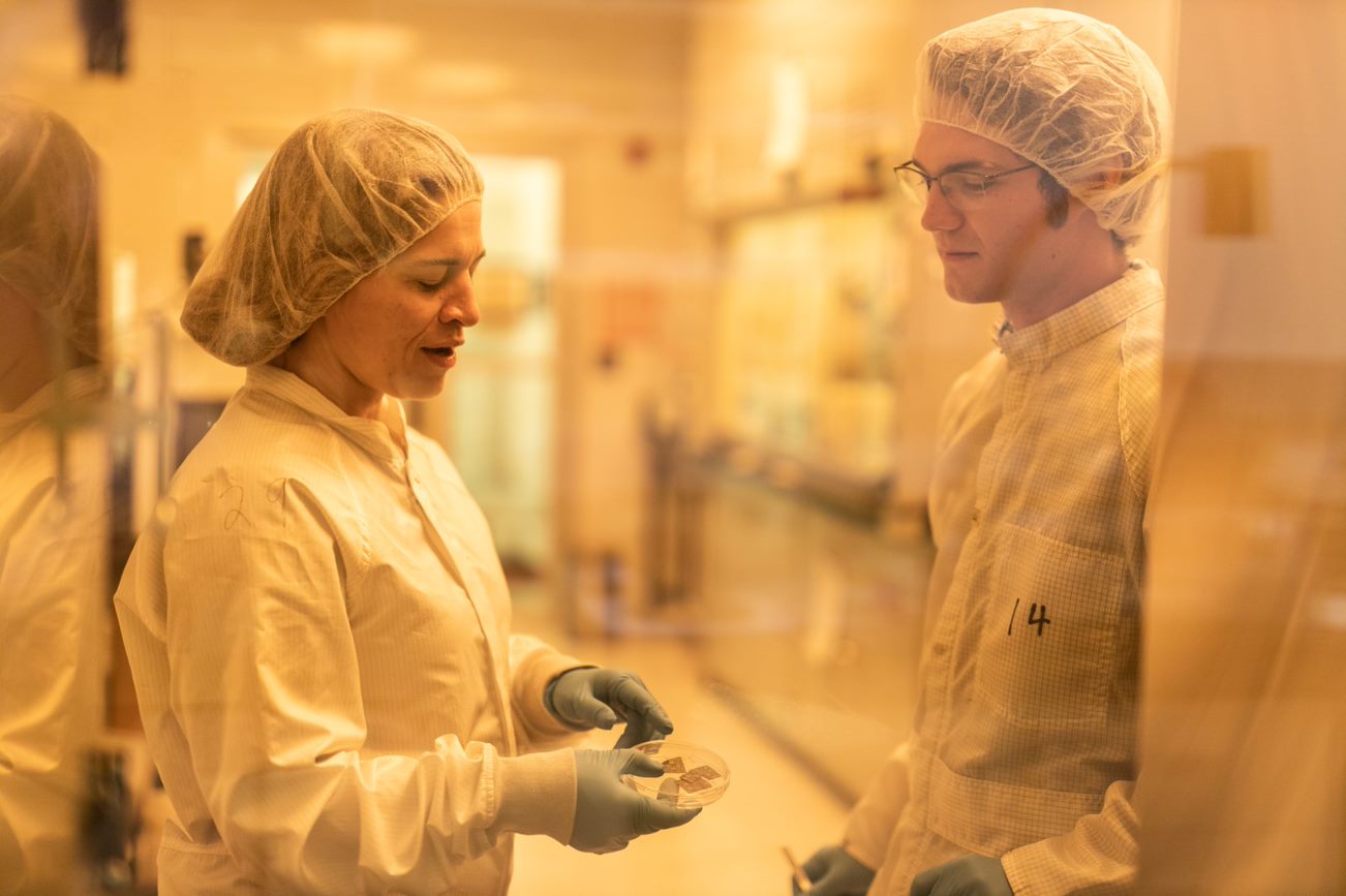 Professor Hinzer and a graduate student in gowns and hair nets looking at a sample in a petri dish in the clean room