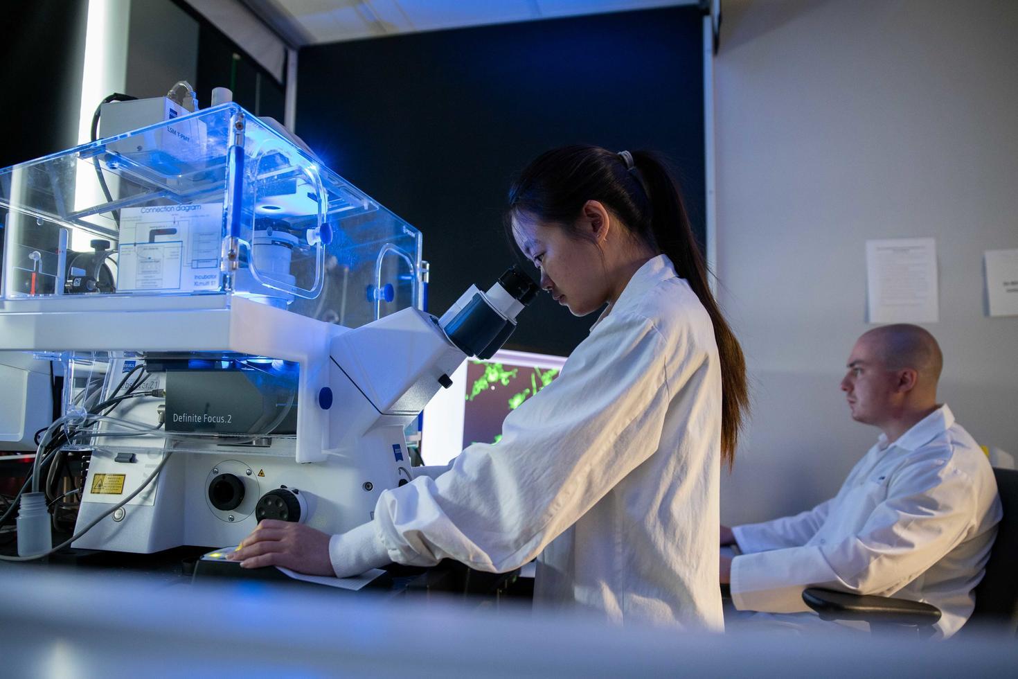 A woman looking through a microscope at the Faculty of Science with a colleague working in the background.