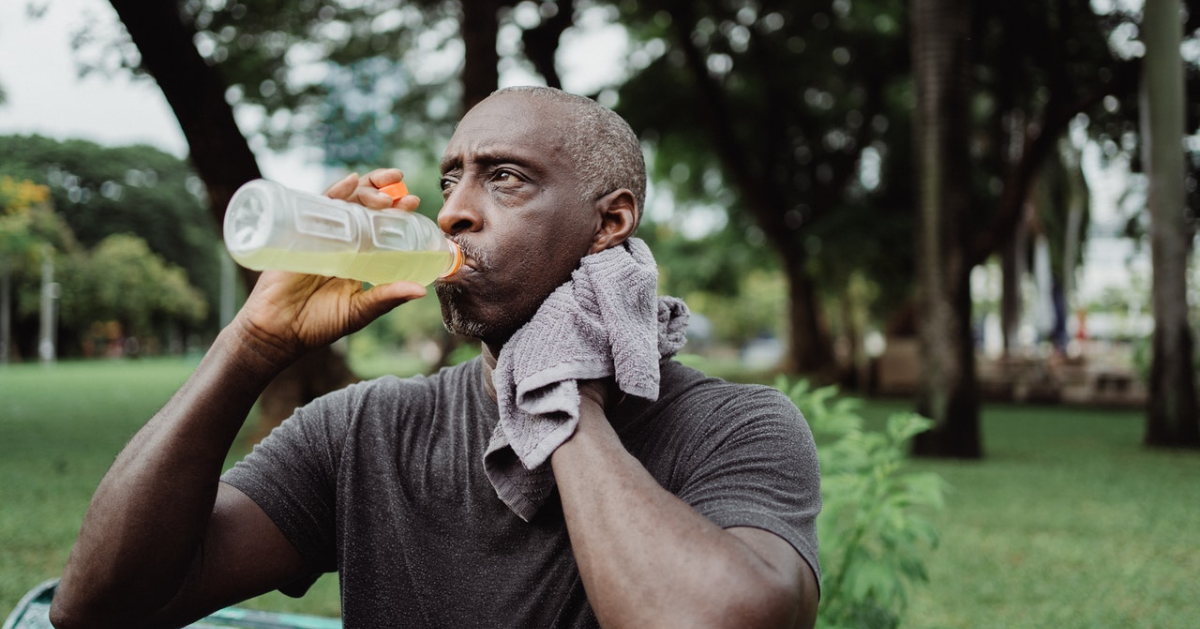 An elderly man sponging his neck with a towel while drinking a sports drink in a bottle