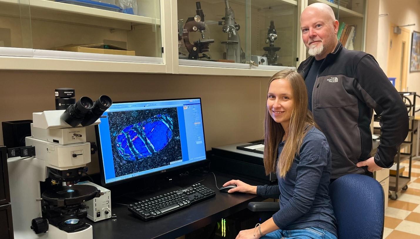 Jennifer Spalding, étudiante en doctorat, est assise à une table de laboratoire devant un ordinateur et le professeur David Schneider se tient à sa droite. 