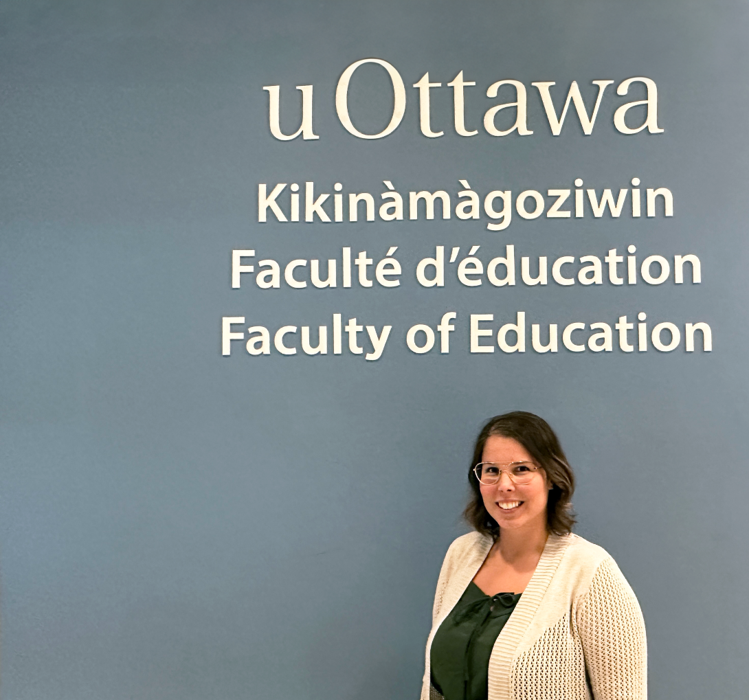 Allison Gagné devant le mur logo de la Faculté avec l'inscription Faculté d'éducation en anglais, en français et en Annishinabe