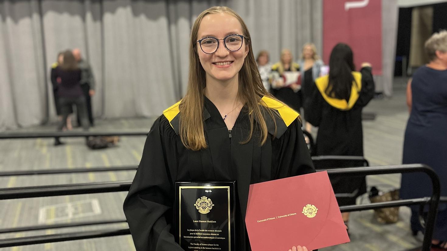 Laura Maddison, wearing a black graduation gown with yellow accents, stands in front of a railing in the uOttawa Convocation Hall. 